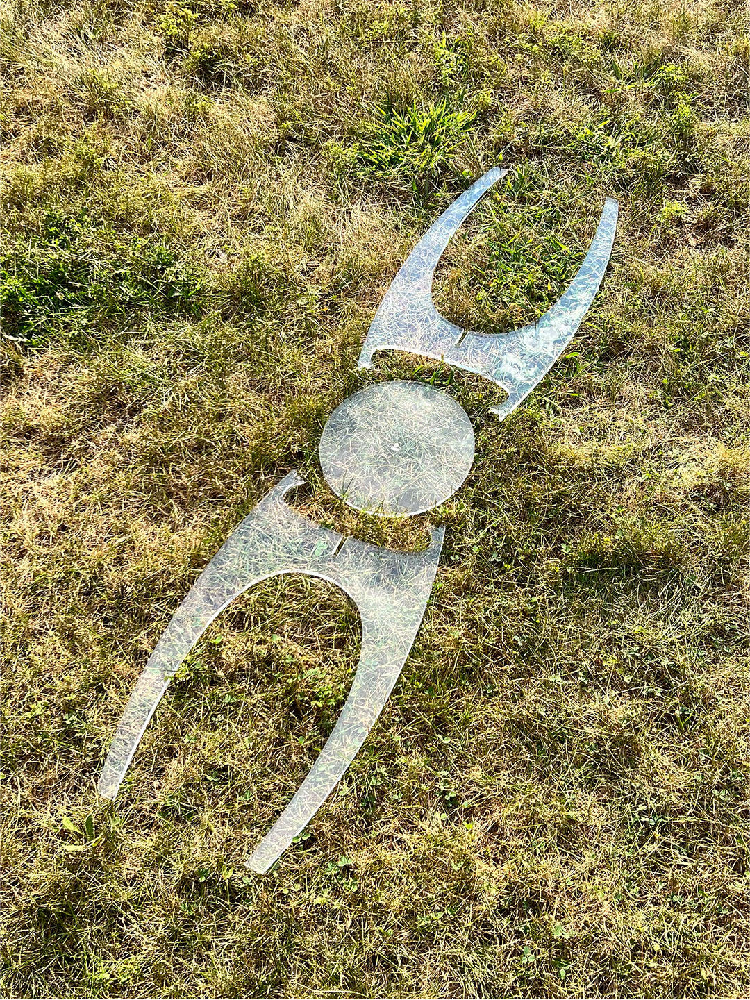 Flat lay of the Aer Table clear acrylic side table disassembled on grass, showing the interlocking curved leg panels and circular tabletop