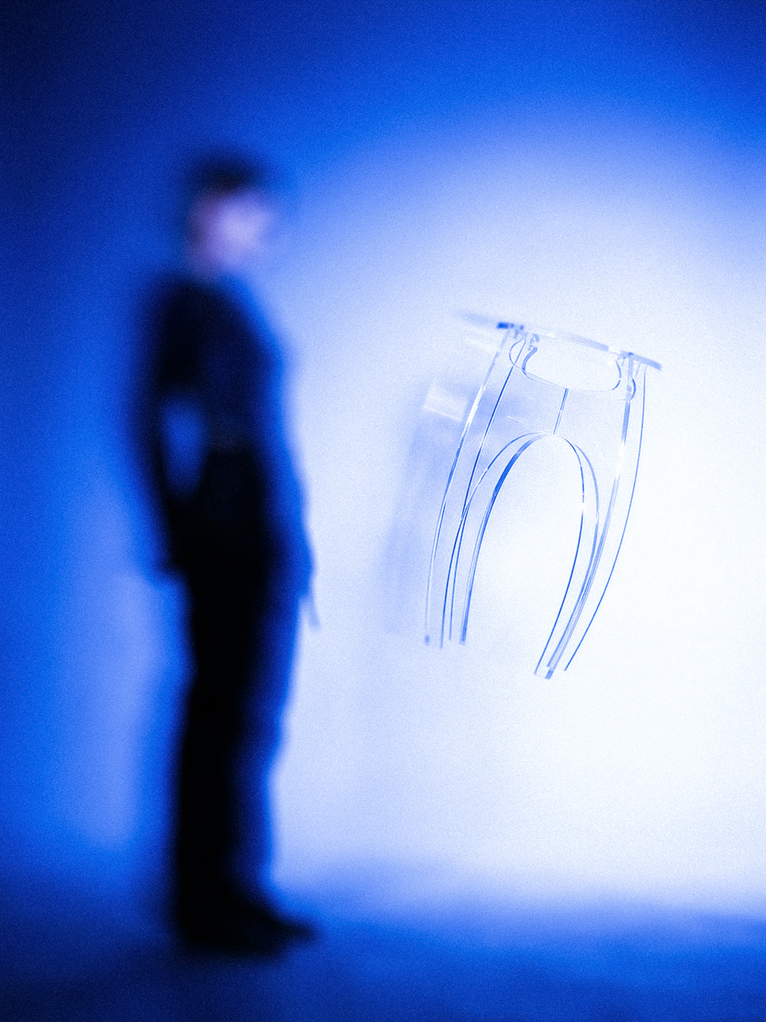 Aer Table clear acrylic side table suspended in a blue-lit environment with a blurred human figure in the background, conveying scale and sculptural presence