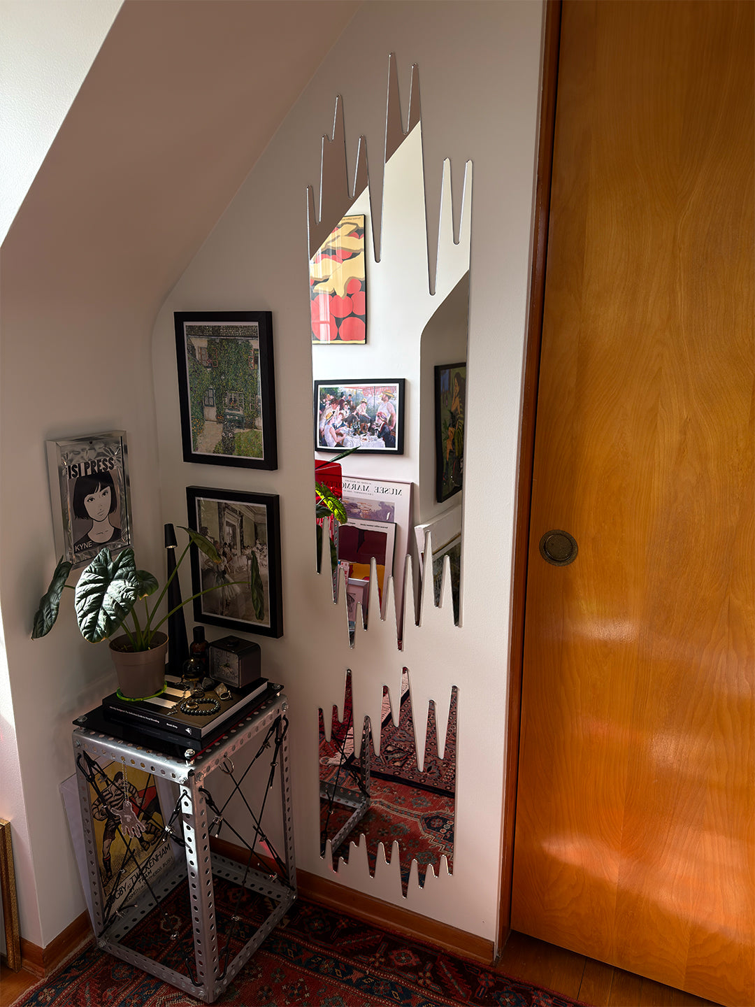 A tall, narrow wall mirror with a jagged, mountain-like silhouette edge is mounted on a slanted white wall in a home interior. The mirror reflects a small shelf area with framed art and books, and the room includes a wooden door, a small metal side table with plants and books, and a patterned rug.