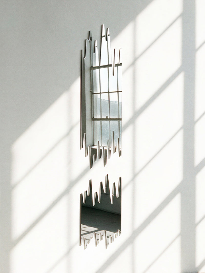An abstract, asymmetrical wall mirror with a metallic silver frame mounted on a white wall, casting soft window shadow patterns across its reflective surface.