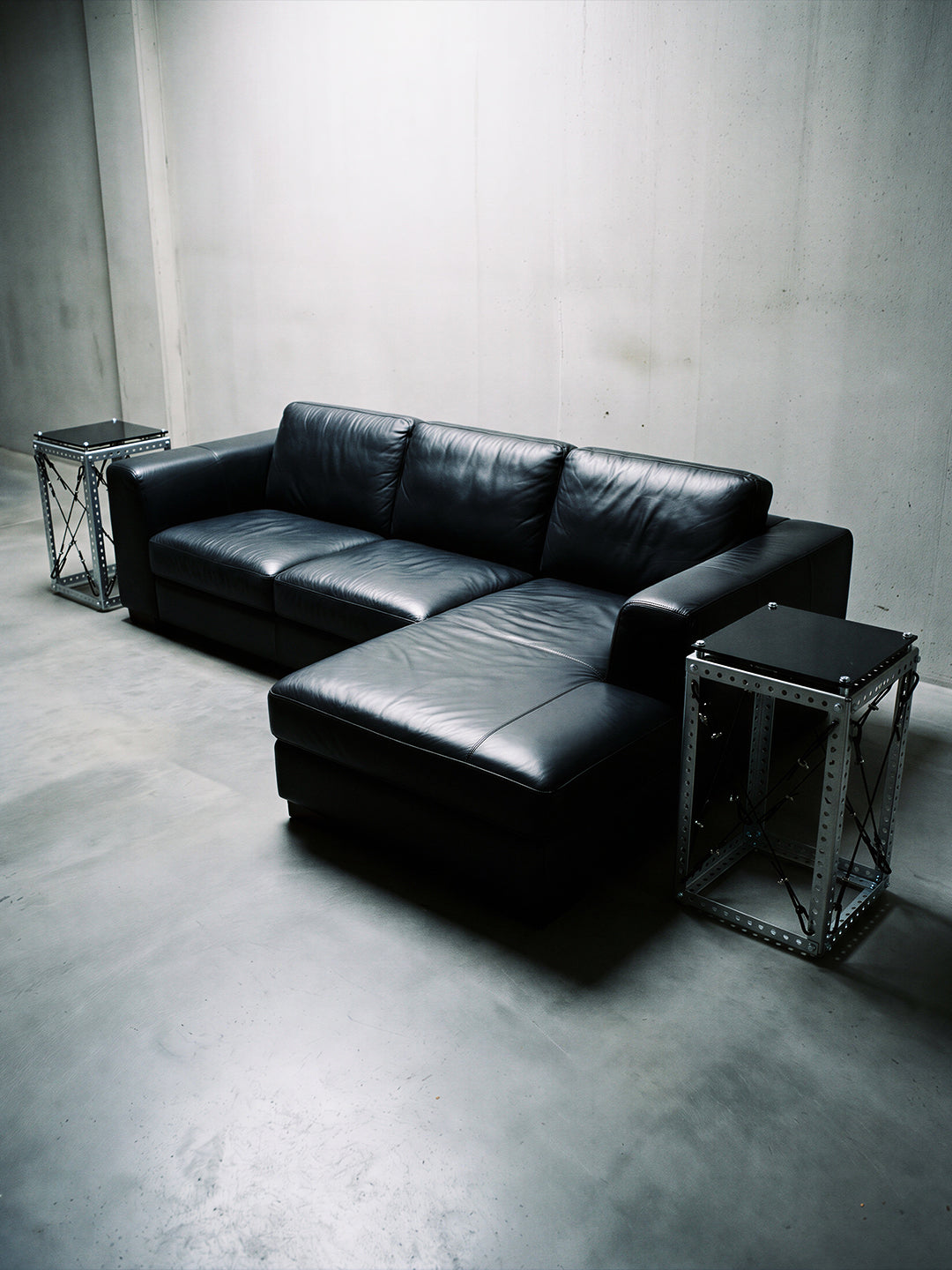 A black leather sectional sofa with a chaise lounge, styled in a minimalist concrete room with two industrial side tables.