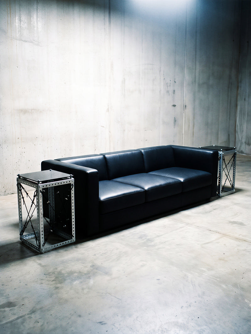 A black leather three-seater sofa with industrial-style metal side tables, set in a concrete warehouse environment.