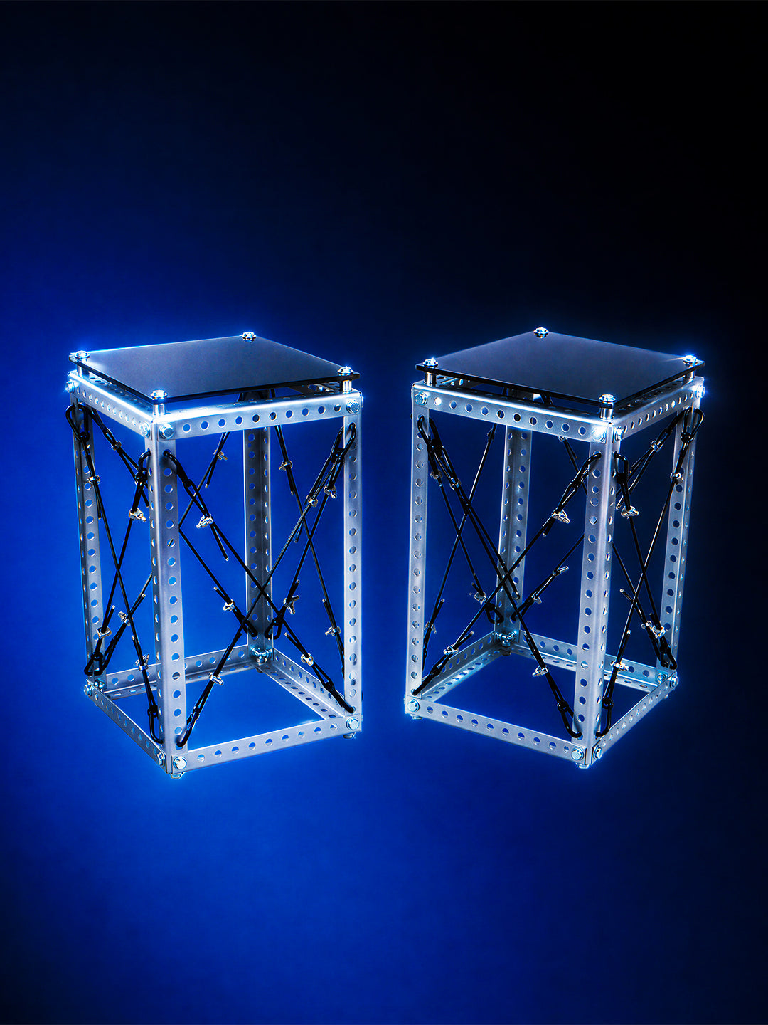 Two modern geometric side tables with clear acrylic panels, silver metal frames, and black crisscross support rods, illuminated with bright blue LED lighting against a dark blue background.